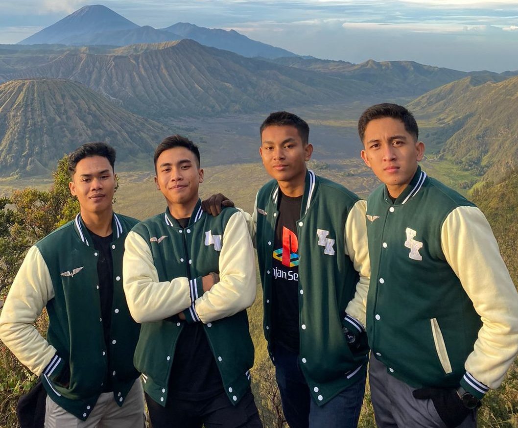 Empat pemuda berpose di latar belakang pegunungan (kemungkinan Gunung Bromo) mengenakan jaket varsity berwarna hijau tua (dark green) dan krem/putih (broken white) dengan aksen logo bordir. Jaket varsity ini sangat populer untuk seragam komunitas, diproduksi oleh konveksi jaket jogja yang juga melayani bordir jogja berkualitas.