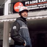 Pria pekerja proyek (engineer/tambang) menggunakan jaket lapangan outdoor berwarna navy/hitam dengan resleting dan bordir logo Adhi dan nama 'A. Syarifudin' di dada, serta helm safety putih, berdiri di area proyek/konstruksi. Jaket ini diproduksi oleh konveksi jogja terbaik, sangat cocok untuk konveksi tambang dan konveksi baju proyek.