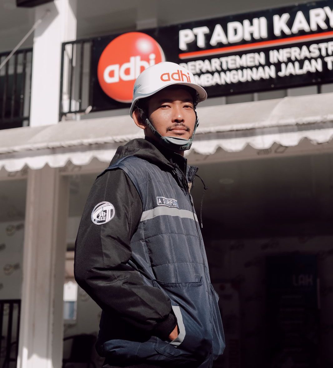 Pria pekerja proyek (engineer/tambang) menggunakan jaket lapangan outdoor berwarna navy/hitam dengan resleting dan bordir logo Adhi dan nama 'A. Syarifudin' di dada, serta helm safety putih, berdiri di area proyek/konstruksi. Jaket ini diproduksi oleh konveksi jogja terbaik, sangat cocok untuk konveksi tambang dan konveksi baju proyek.